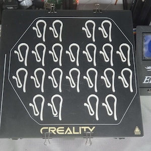 Puede incluir: Una base de impresora 3D negra con clips blancos en forma de U dispuestos en filas. La palabra "CREALITY" está impresa en blanco en la parte inferior. Los clips metálicos aseguran la base. Un panel de control de la impresora 3D es visible a la derecha.