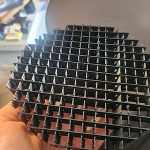 May include: A black metal grid with a geometric, repeating pattern of rectangular openings. The grid is held in a hand, with a blurred background. The grid appears to be a component of a larger structure.
