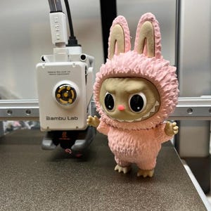 May include: A pink, furry figurine of a creature with bunny ears and a toothy grin. The figure stands on a textured surface near a 3D printer with the text "Bambu Lab". The figurine has a light brown face and black eyes.