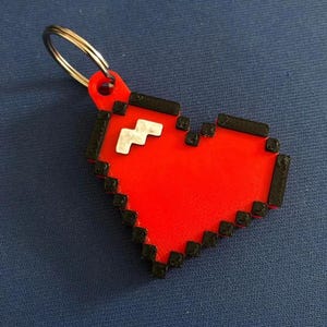 Könnte beinhalten: Ein pixeliger roter Herz-Schlüsselanhänger mit schwarzen Umrissen und einem silbernen Schlüsselring. Das Herz hat ein weißes Zickzack-Design in der Mitte. Ein lustiges Accessoire.