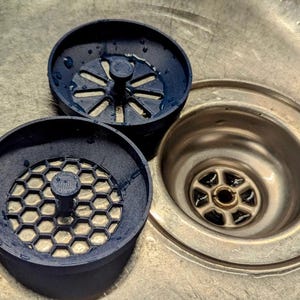 May include: Three dark blue sink strainers with different patterned grates sit next to a stainless steel sink drain. One strainer has a starburst pattern, and the other has a honeycomb pattern. The sink drain is circular.