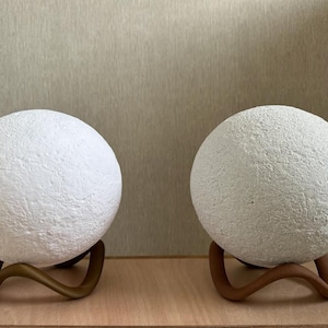May include: Two white, textured sphere lamps with a moon-like appearance. Each lamp sits on a curved, bronze-colored stand. The lamps are displayed on a light-colored wooden surface against a neutral background.