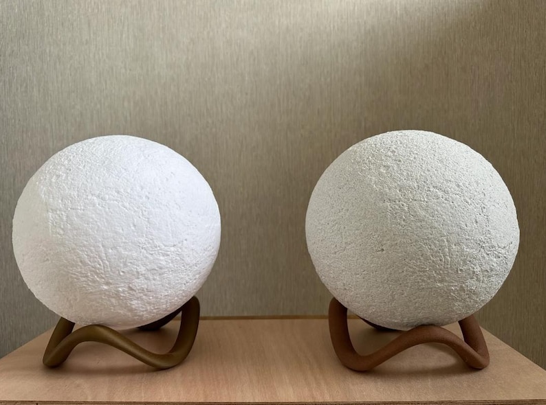 May include: Two white, textured sphere lamps with a moon-like appearance. Each lamp sits on a curved, bronze-colored stand. The lamps are displayed on a light-colored wooden surface against a neutral background.