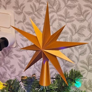 May include: A gold-colored star tree topper with multiple points. The star is mounted on a cone-shaped base and is placed on top of a Christmas tree with green needles. The star has a layered design and is made of a metallic material.