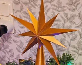 Puede incluir: Una estrella dorada para la parte superior del árbol de Navidad con múltiples puntas. La estrella está montada sobre una base en forma de cono y se coloca encima de un árbol de Navidad con agujas verdes. La estrella tiene un diseño en capas y es de metal.