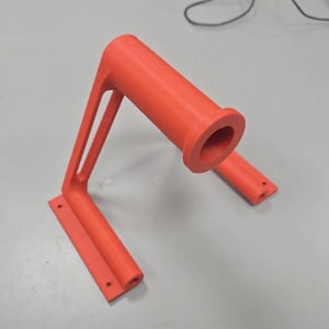 Puede incluir: Un soporte de color naranja brillante, impreso en 3D, con un asa cil&iacute;ndrica y una base rectangular. El soporte tiene un dise&ntilde;o en &aacute;ngulo recto con dos puntales de soporte. La base tiene tres agujeros para el montaje. El asa tiene una gran abertura.
