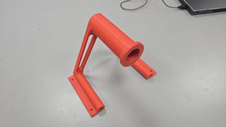 Puede incluir: Un soporte de color naranja brillante, impreso en 3D, con un asa cil&iacute;ndrica y una base rectangular. El soporte tiene un dise&ntilde;o en &aacute;ngulo recto con dos puntales de soporte. La base tiene tres agujeros para el montaje. El asa tiene una gran abertura.