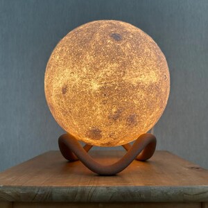 May include: A glowing, spherical moon lamp with a textured surface, resting on a curved, brown stand. The lamp emits a warm, inviting light, creating a celestial ambiance. The lamp is placed on a wooden table.