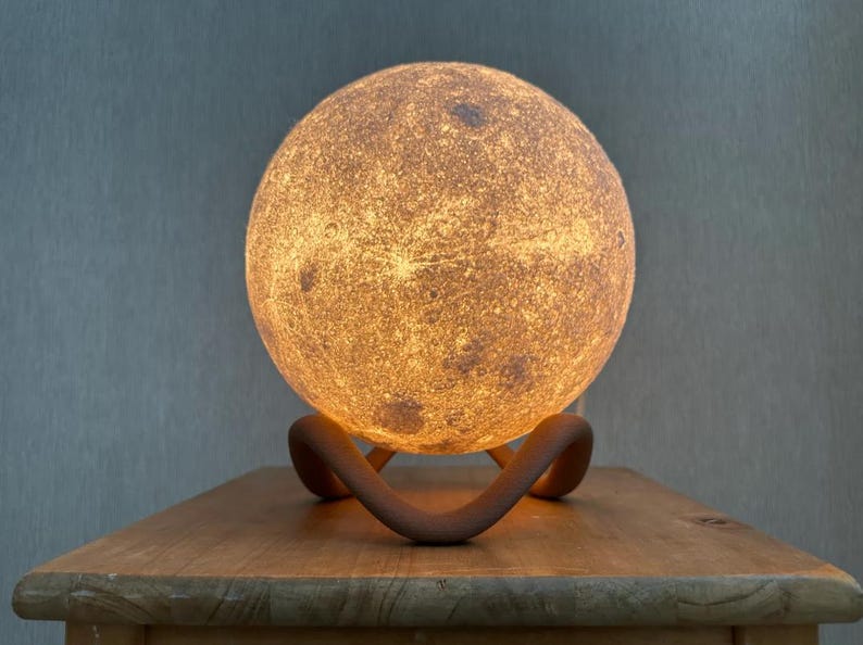 May include: A glowing, spherical moon lamp with a textured surface, resting on a curved, brown stand. The lamp emits a warm, inviting light, creating a celestial ambiance. The lamp is placed on a wooden table.