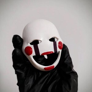 以下が含まれることがあります： 赤い丸い頬と赤い笑顔が特徴の白いマスク。 マスクには黒い眼窩と紫の垂直線があります。 マスクは黒い手袋を着用した人が持っています。