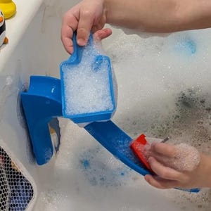 May include: A blue plastic bath toy with a slide and a scoop filled with bubbles. A red sponge is being used on the slide. The toy is in a bathtub filled with bubbles. A yellow and black penguin toy is visible.