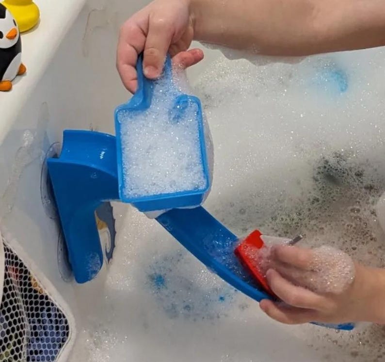 May include: A blue plastic bath toy with a slide and a scoop filled with bubbles. A red sponge is being used on the slide. The toy is in a bathtub filled with bubbles. A yellow and black penguin toy is visible.