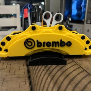 May include: A yellow desk organizer shaped like a Brembo brake caliper, holding pens, scissors, and a utility knife. The organizer has the Brembo logo in black and is designed to hold office supplies.