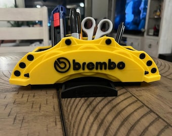 May include: A yellow desk organizer shaped like a Brembo brake caliper, holding pens, scissors, and a utility knife. The organizer has the Brembo logo in black and is designed to hold office supplies.