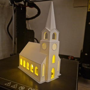 Peut inclure: Modèle d'église blanc imprimé en 3D, avec un haut clocher et des fenêtres en arc. Le modèle est éclairé de l'intérieur, projetant une lueur jaune chaude à travers les ouvertures. L'église est posée sur une surface noire.