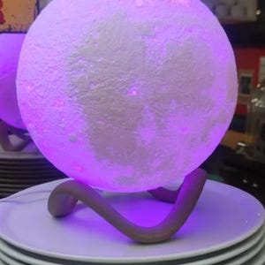 May include: A glowing, spherical moon lamp with a textured surface, emitting a soft purple light. The lamp rests on a curved, brown stand, placed atop a stack of white plates. The lamp is a decorative lighting fixture.