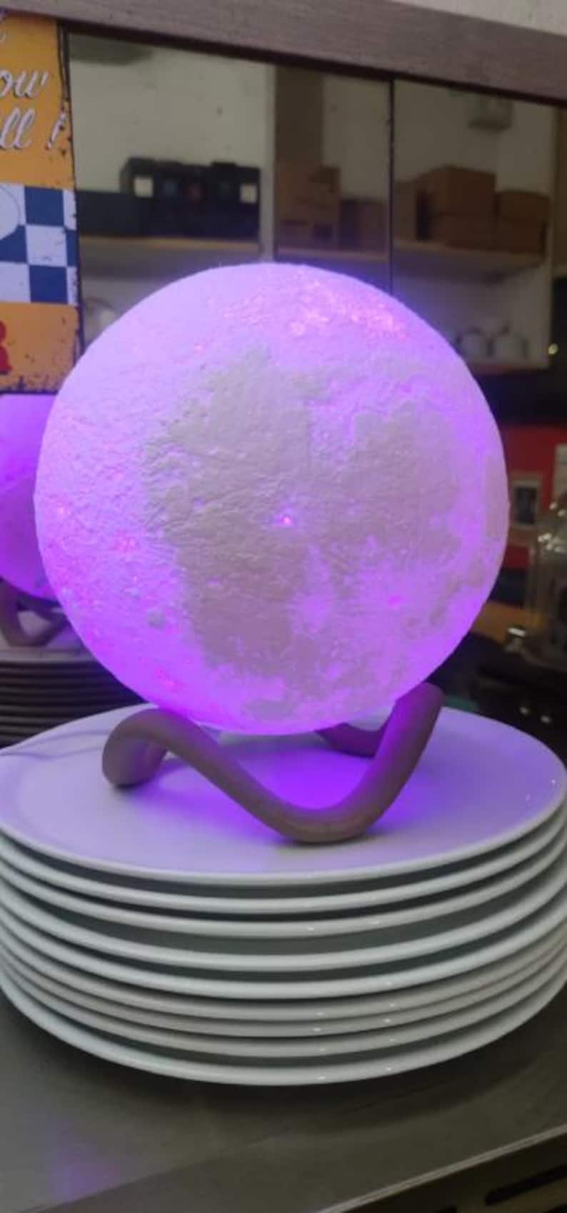 May include: A glowing, spherical moon lamp with a textured surface, emitting a soft purple light. The lamp rests on a curved, brown stand, placed atop a stack of white plates. The lamp is a decorative lighting fixture.