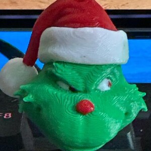 May include: A green Grinch-themed Christmas ornament with a red Santa hat and a red nose. The ornament has a loop for hanging and is designed to resemble the Grinch character from the popular holiday story.