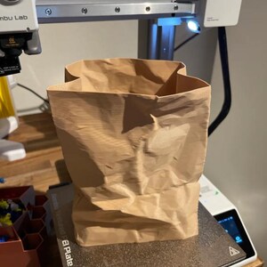 May include: A crumpled brown paper bag sits on a 3D printer bed. The bag is rectangular with a wide opening and appears to be made of a thick paper material. The 3D printer has a silver frame and a small light.