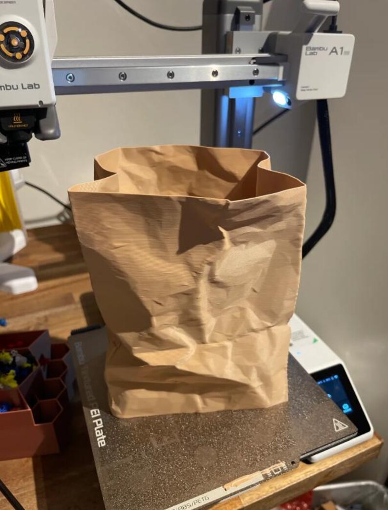 May include: A crumpled brown paper bag sits on a 3D printer bed. The bag is rectangular with a wide opening and appears to be made of a thick paper material. The 3D printer has a silver frame and a small light.