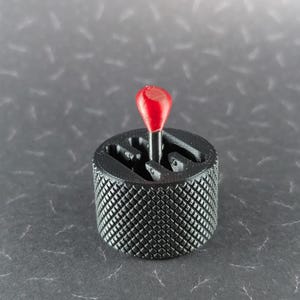 May include: A black gear shift knob with a red top. The knob has a diamond pattern texture and a gear shift pattern. The red top is a rounded shape.