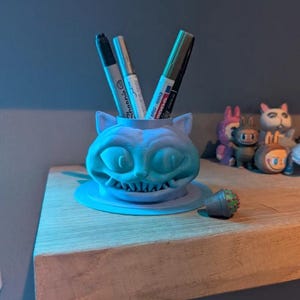 Peut inclure: Un porte-stylos bleu clair imprimé en 3D en forme du Chat du Cheshire. Le support a un large sourire et contient plusieurs stylos et marqueurs. Un petit cactus décoratif est à proximité.