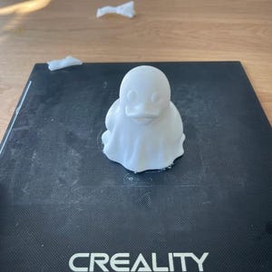 May include: A white 3D-printed duck figurine with a rounded body and a small beak. The duck is sitting on a black surface with the word "CREALITY" printed on it. The background includes a wooden table and some white paper.