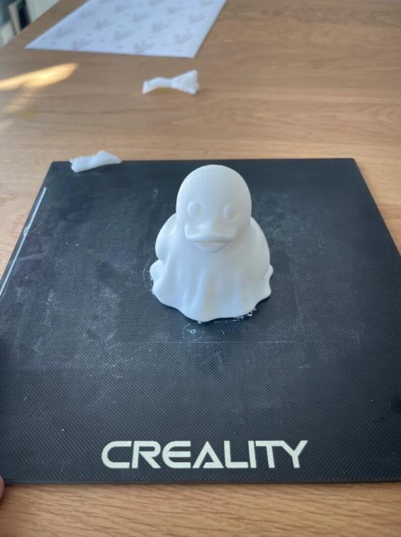 May include: A white 3D-printed duck figurine with a rounded body and a small beak. The duck is sitting on a black surface with the word "CREALITY" printed on it. The background includes a wooden table and some white paper.