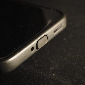 以下が含まれることがあります： 黒いスマートフォンのクローズアップ。電源ボタン、小さな穴、スピーカーグリルが表示され、電話機の端が見えます。電話の画面は暗く、周囲の環境を反射しています。
