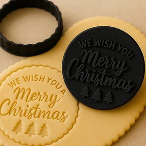 May include: A black cookie stamp and cutter set for Christmas. The stamp reads "We Wish You a Merry Christmas" with three small Christmas trees. A stamped cookie and a black cutter are also visible.