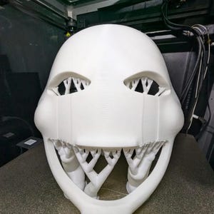 以下が含まれることがあります： 白い、3Dプリントされたマスク。 目の部分と口の部分が開いており、複雑な木の様な構造になっています。小道具やコスチュームの一部である可能性があります。