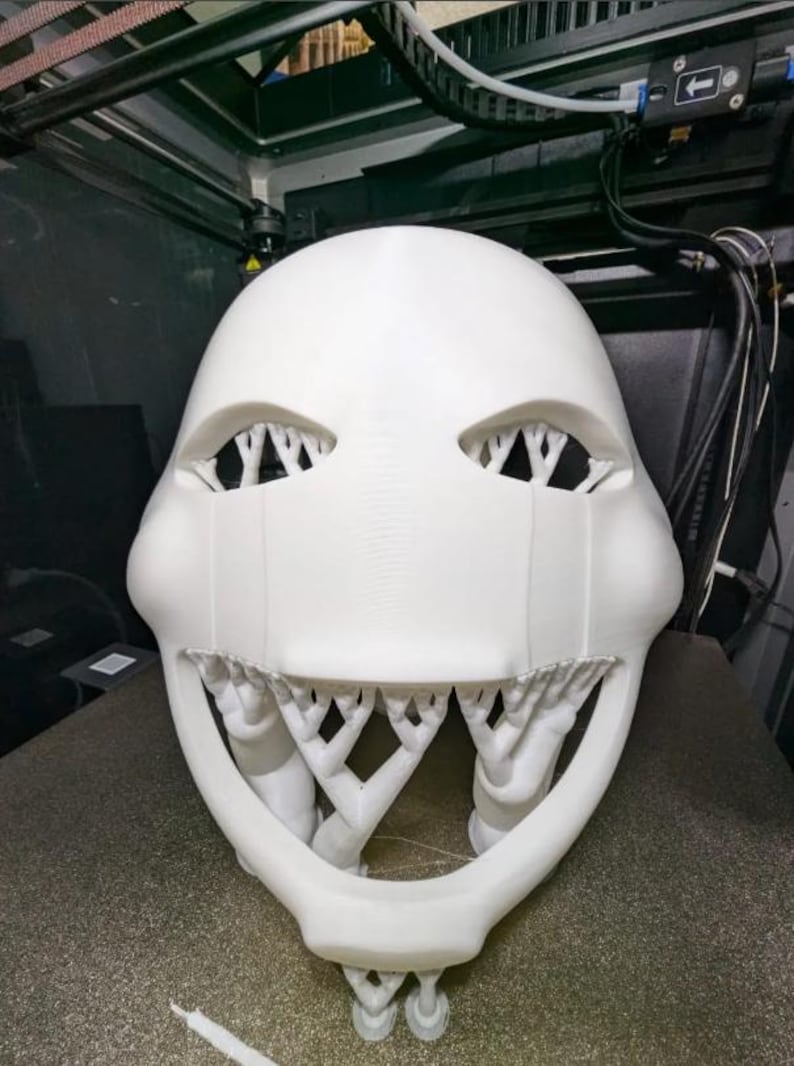 以下が含まれることがあります： 白い、3Dプリントされたマスク。 目の部分と口の部分が開いており、複雑な木の様な構造になっています。小道具やコスチュームの一部である可能性があります。