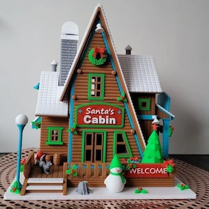 May include: A detailed Christmas-themed cabin with a brown wooden exterior, green trim, and a white roof. The sign reads "Santa's Cabin" in red. A snowman, a Christmas tree, and a welcome sign are in front of the cabin. A small dog statue is on the porch.