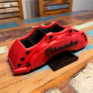 May include: A red desk organizer shaped like a Brembo brake caliper. The organizer has black interior compartments and the Brembo logo on the side. A small black stand is included. The item is on a wooden surface.