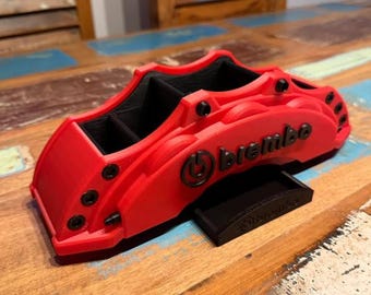 May include: A red desk organizer shaped like a Brembo brake caliper. The organizer has black interior compartments and the Brembo logo on the side. A small black stand is included. The item is on a wooden surface.