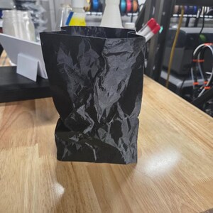 May include: A black, crumpled paper bag stands on a wooden surface. The bag has a rectangular base and a slightly wider top, with a textured, wrinkled appearance. The lighting creates shadows that emphasize the bag's shape.