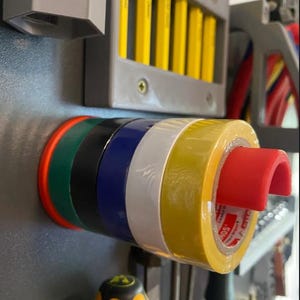 Puede incluir: Primer plano de una colección de rollos de cinta aislante de colores, incluyendo rojo, verde, negro, azul, blanco y amarillo. Un soporte rojo sostiene el rollo amarillo. La imagen también muestra lápices amarillos y herramientas.