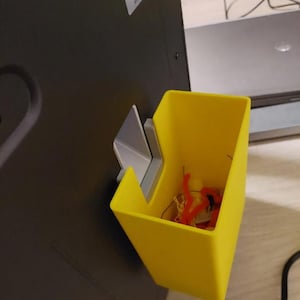 Könnte beinhalten: Ein leuchtend gelber, an der Wand befestigter Aufbewahrungsbehälter mit einer silbernen Halterung. Der Behälter enthält rote und gelbe Bastelmaterialien. Der Behälter ist an einer dunkelgrauen Oberfläche befestigt.
