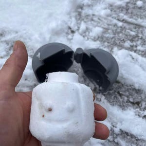 Può includere: Una testa bianca scolpita nella neve, in stile Lego, con un volto sorridente, tenuta in mano. Uno stampo grigio sferico è aperto sullo sfondo, su una superficie innevata. Un divertente lavoretto a tema invernale.