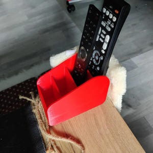 May include: A vibrant red remote control tidy with three sections. Two black remote controls are stored within. The tidy is positioned on a wooden surface.