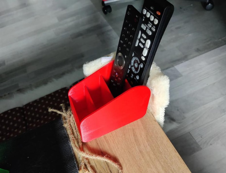 May include: A vibrant red remote control tidy with three sections. Two black remote controls are stored within. The tidy is positioned on a wooden surface.