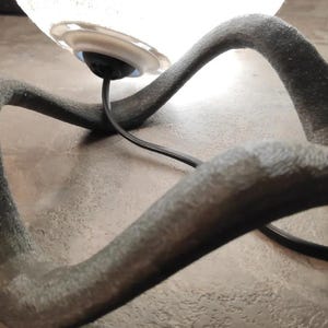 May include: A modern table lamp with a clear glass shade and a dark gray, curved base. The base has a textured surface and a black power cord. The lamp is designed with an artistic, flowing shape.