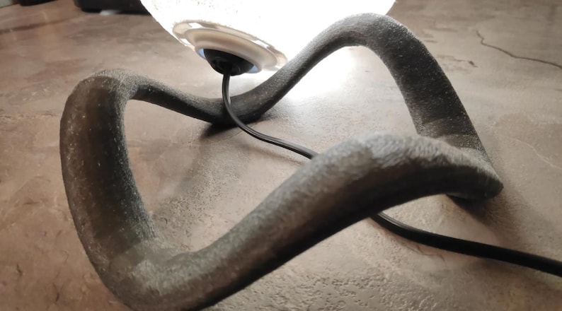 May include: A modern table lamp with a clear glass shade and a dark gray, curved base. The base has a textured surface and a black power cord. The lamp is designed with an artistic, flowing shape.