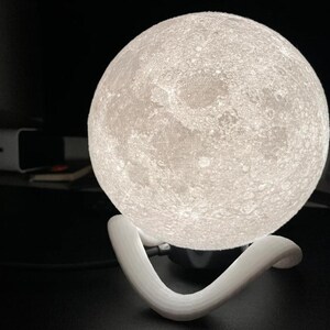 May include: A glowing, spherical moon lamp with a textured surface, resting on a white, curved stand. The lamp emits a soft, warm light, creating a celestial ambiance. The lamp is a decorative lighting fixture.