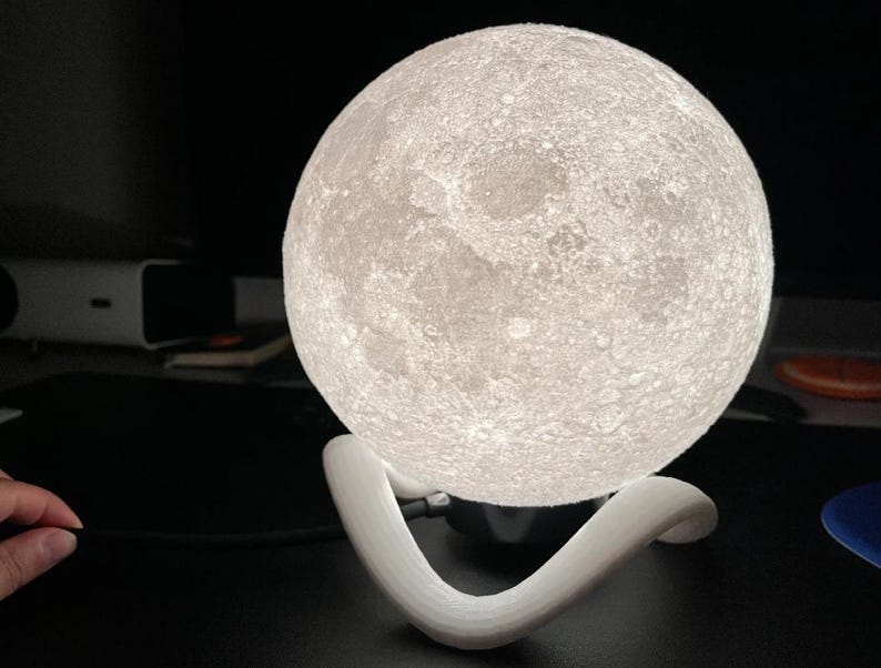 May include: A glowing, spherical moon lamp with a textured surface, resting on a white, curved stand. The lamp emits a soft, warm light, creating a celestial ambiance. The lamp is a decorative lighting fixture.