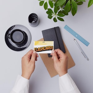 Puede incluir: Una persona sostiene una tarjeta con la palabra "FLASHCARDS" en naranja y negro. La tarjeta está sobre un escritorio con una taza y un platillo negros, un cuaderno, una regla y un bolígrafo.