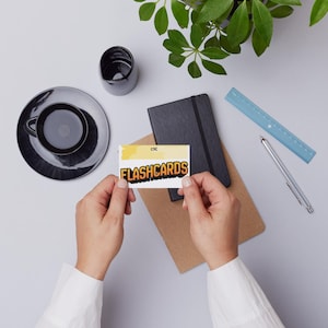 Puede incluir: Vista aérea de un espacio de trabajo con una taza y un platillo negros, un cuaderno negro y una regla. Una persona sostiene una tarjeta con la palabra "FLASHCARDS". También se ven un bolígrafo y una planta pequeña.