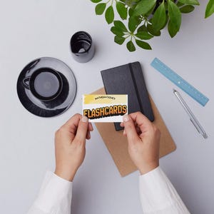 Peut inclure: Vue de dessus d'un espace de travail avec une tasse et une soucoupe noires, un carnet noir, une règle bleue et un stylo. Une personne tient une carte mémoire avec le mot "FLASHCARDS". Une petite plante est en haut à droite.