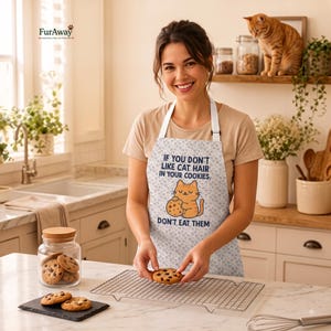 Peut inclure: Un tablier blanc orné d'un motif répétitif de petites empreintes de pattes et d'un dessin central représentant un chat orange tenant un cookie aux pépites de chocolat. Le texte indique : IF YOU DON'T LIKE CAT HAIR IN YOUR COOKIES. DON'T EAT THEM.