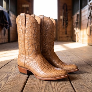 Puede incluir: Un par de botas vaqueras de color tostado con un patrón de textura de piel de cocodrilo, punta puntiaguda, tacón de madera apilado y costuras decorativas en la caña alta. Las botas tienen un acabado claro y una silueta occidental clásica.